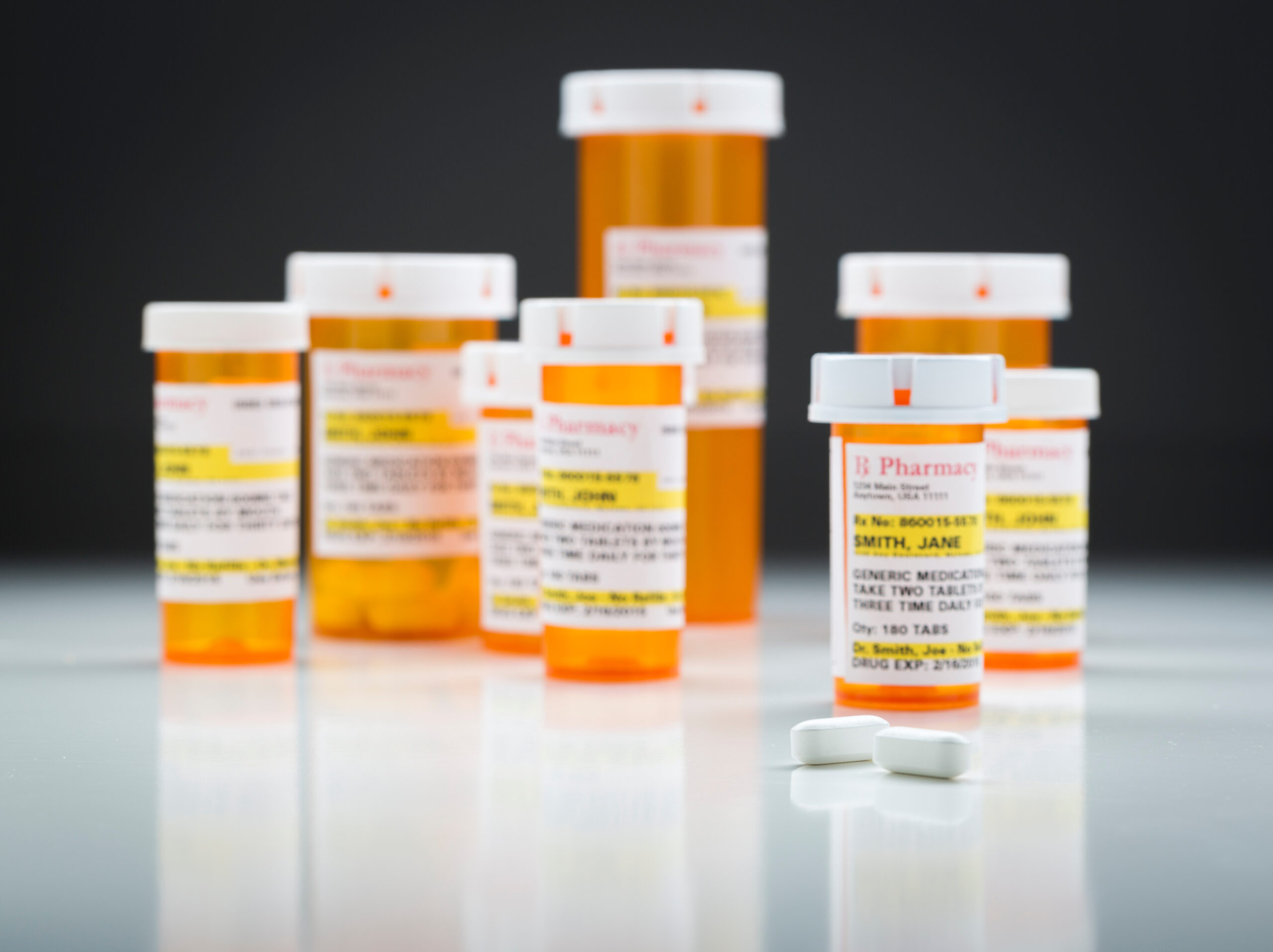 Variety of Non-Proprietary Medicine Bottles and Large Pills on Reflective Surface With Grey Background.