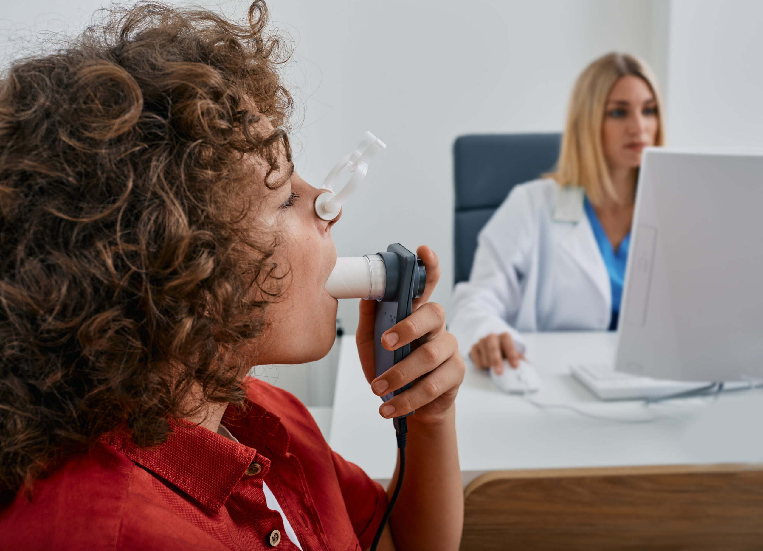 Male child during breathing spirometry and pulmonary function test using medical spirometer with doctor looking at test results on monitor