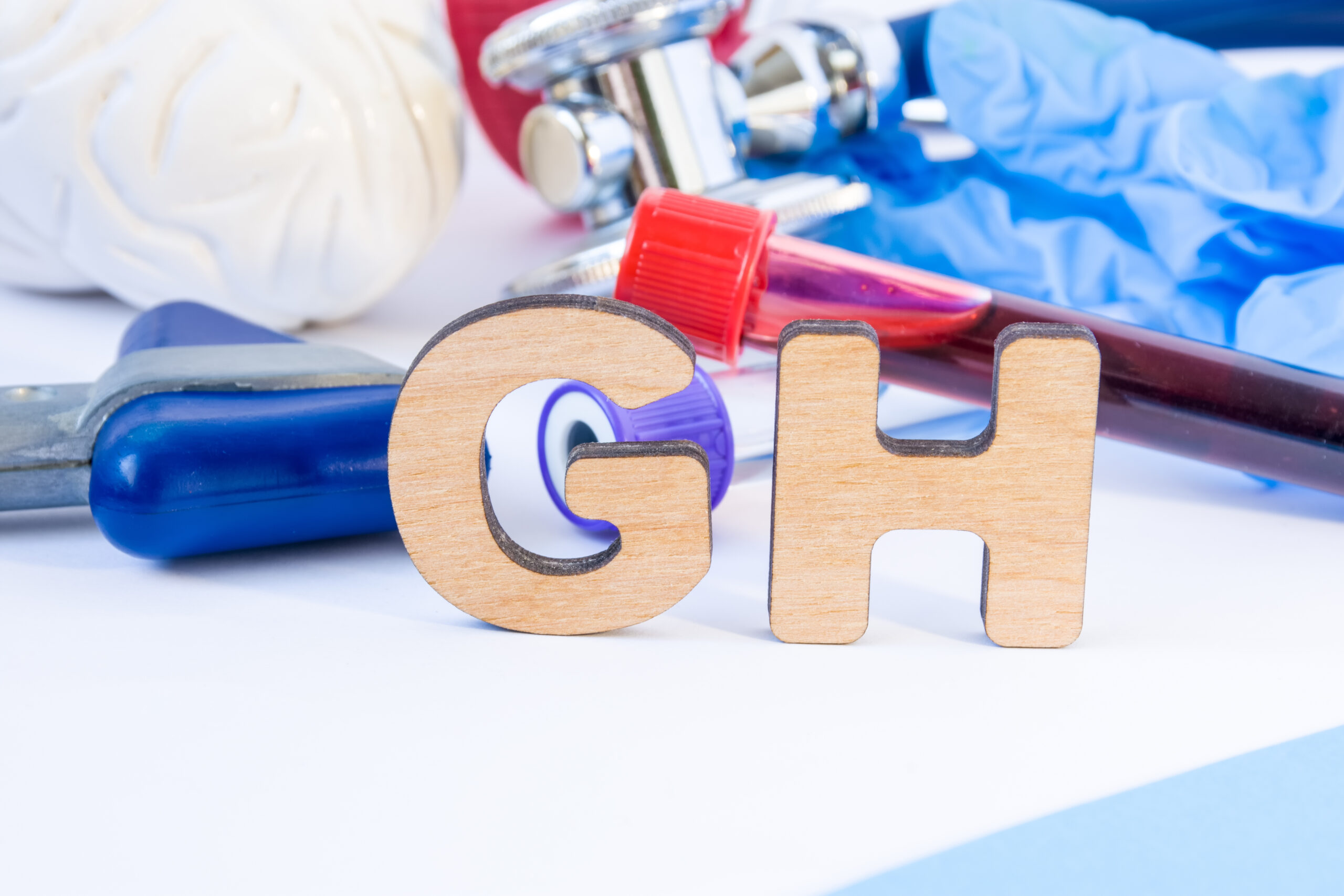 GH abbreviation or acronym in foreground in laboratory scientific or medical practice meaning growth hormone or somatotropin, with model of brain, neurological hammer laboratory test tubes stethoscope